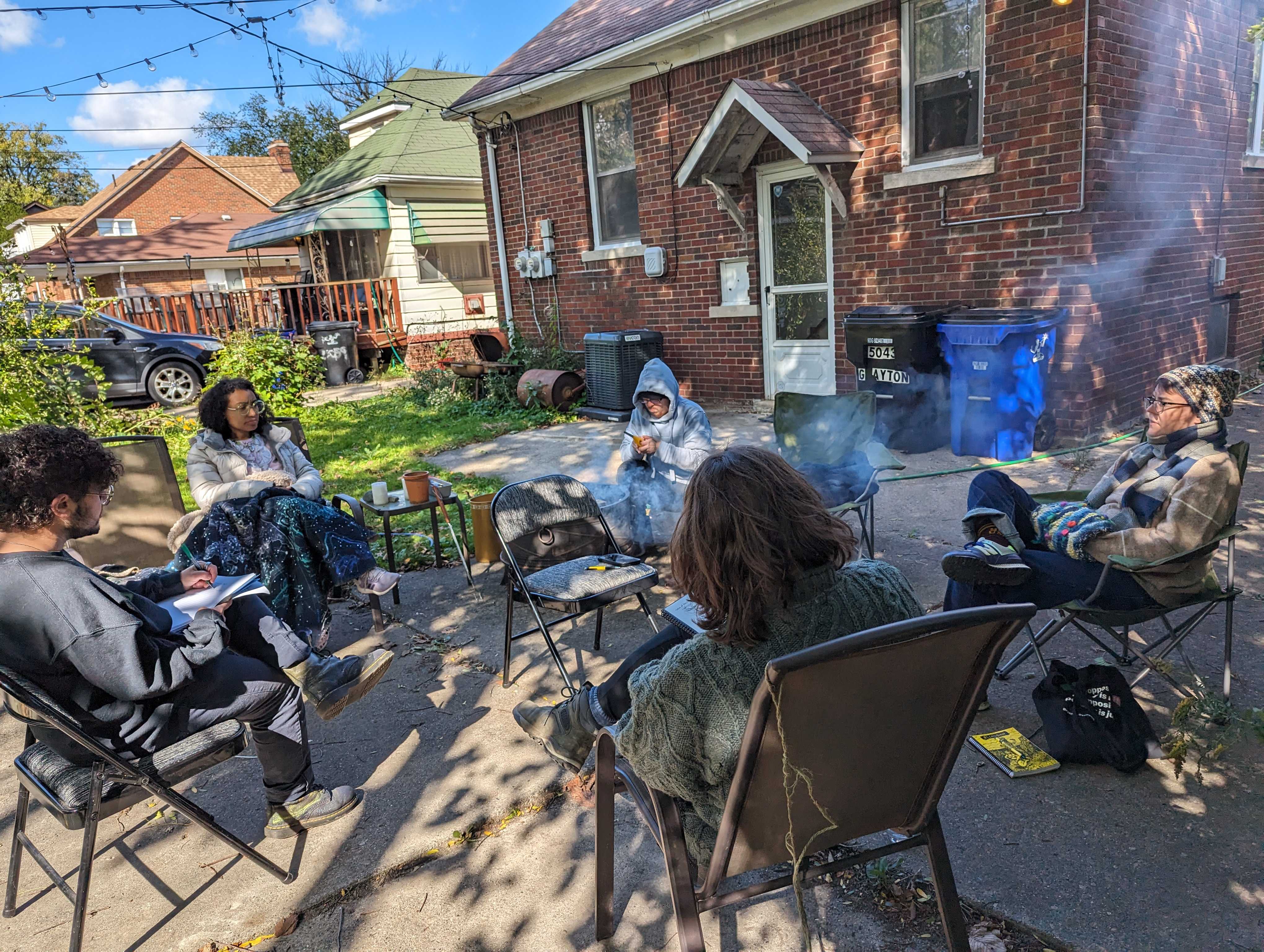 A photo of Detroit Peer Respite members sitting around a fire on folding chairs. Most of them are taking notes.
