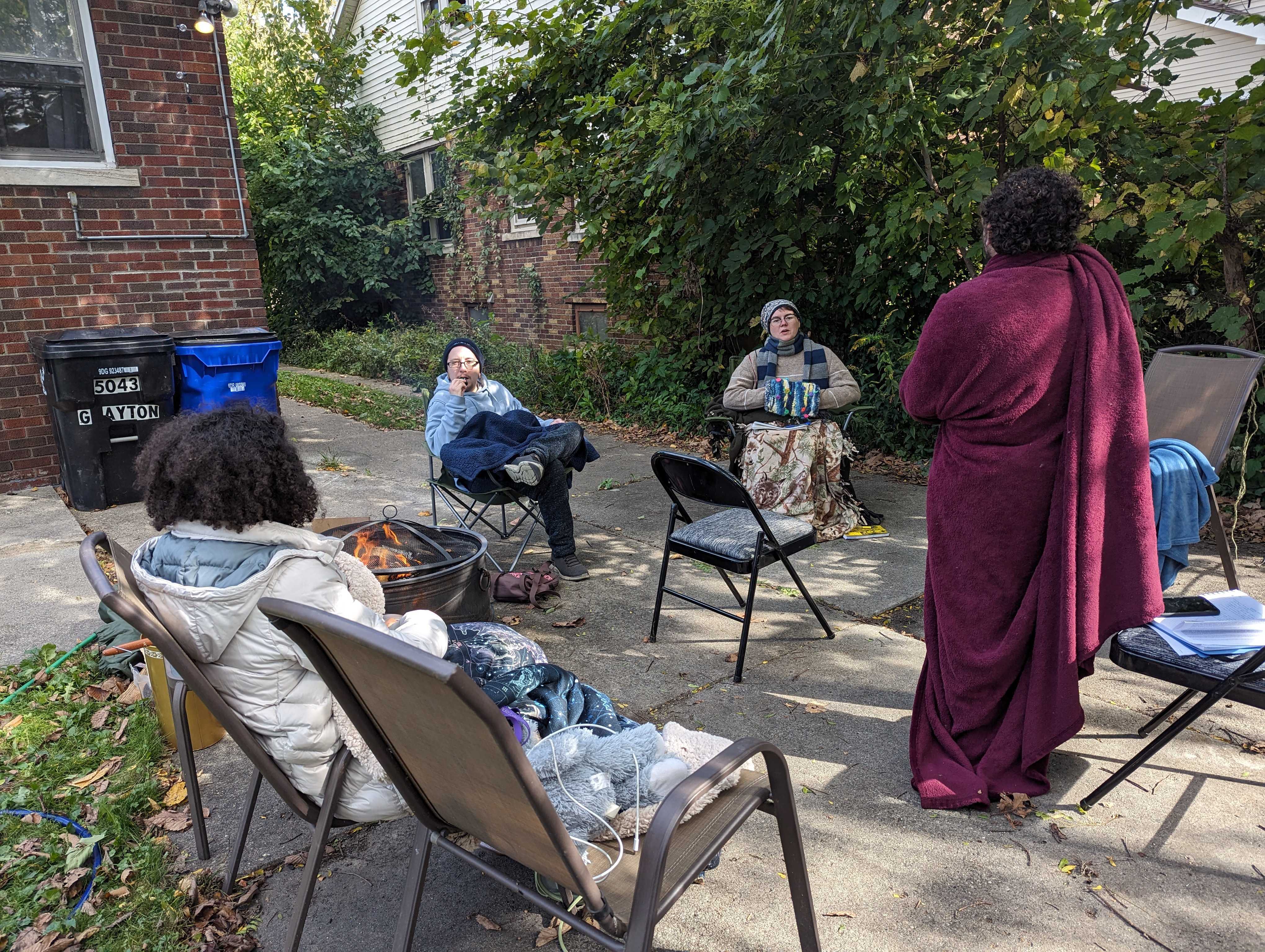 A photo of Detroit Peer Respite members sitting around a fire on folding chairs. Grey is standing up, draped dramatically in a fuzzy red blanket.