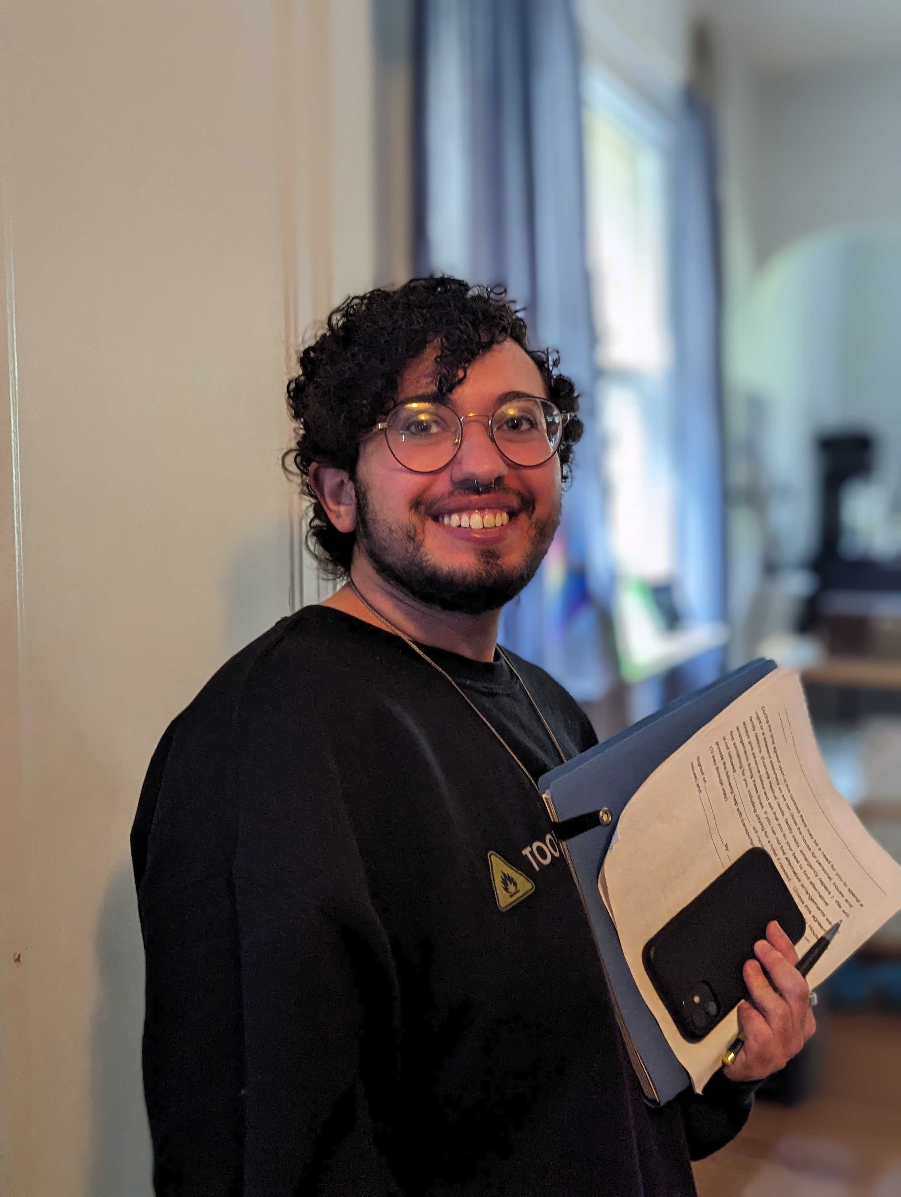 A photo of Grey, a member of Detroit Peer Respite, looking at the camera and smiling awkwardly. He holds a notebook in one hand.