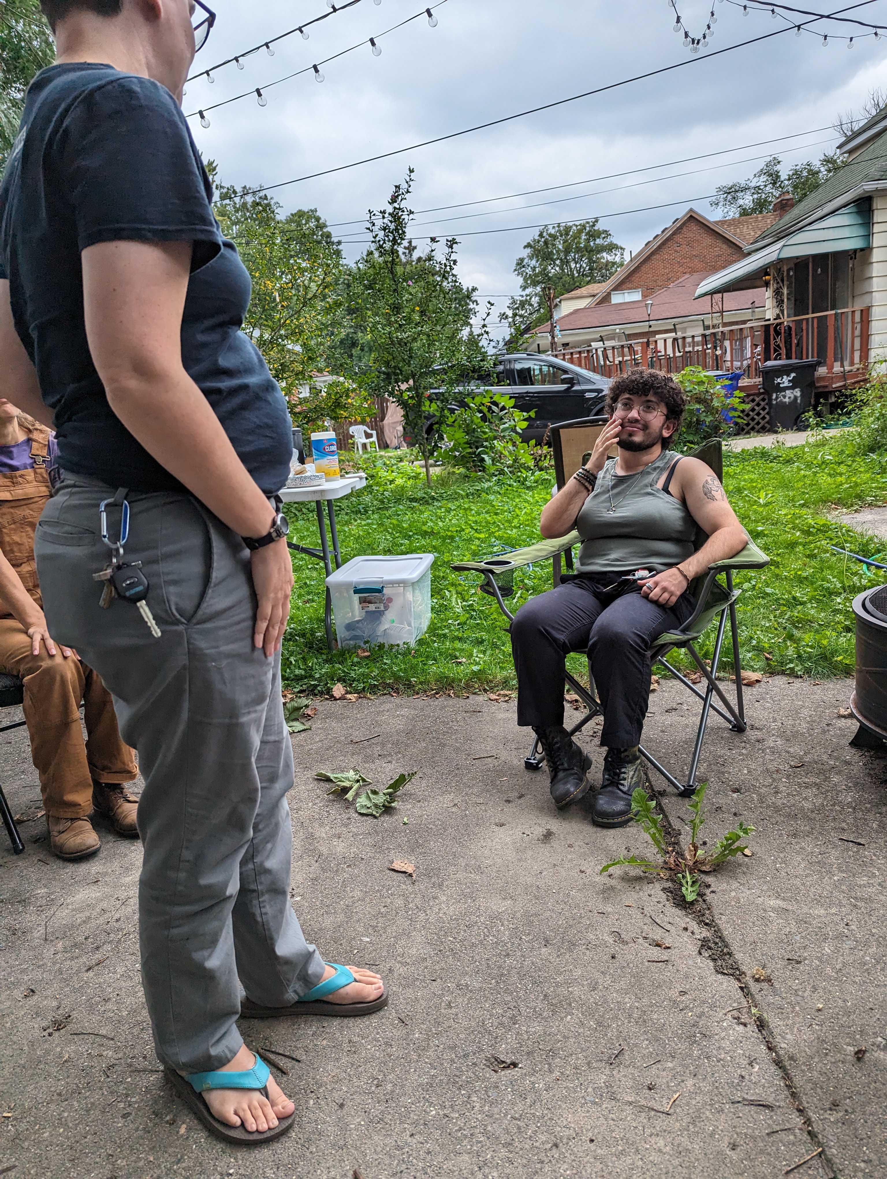 A photo of Grey and Meg, two Detroit Peer Respite members. Grey is sitting in a folding chair and looking up at Meg, who stands in front of him; both are smiling.