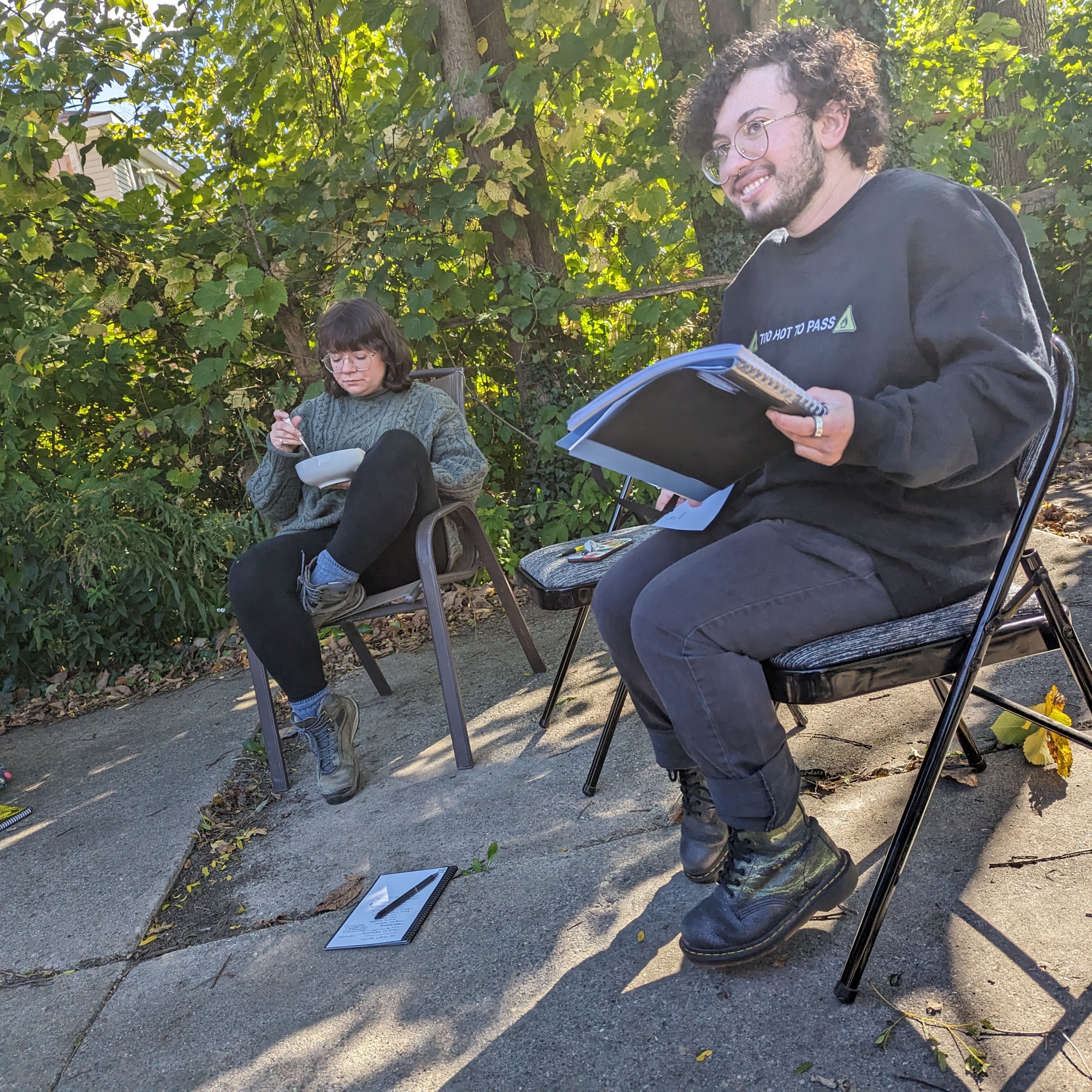 A photo of Grey and Erin, two members of Detroit Peer Respite, sitting on folding chairs. Grey is writing in a notebook while Erin eats from a bowl.