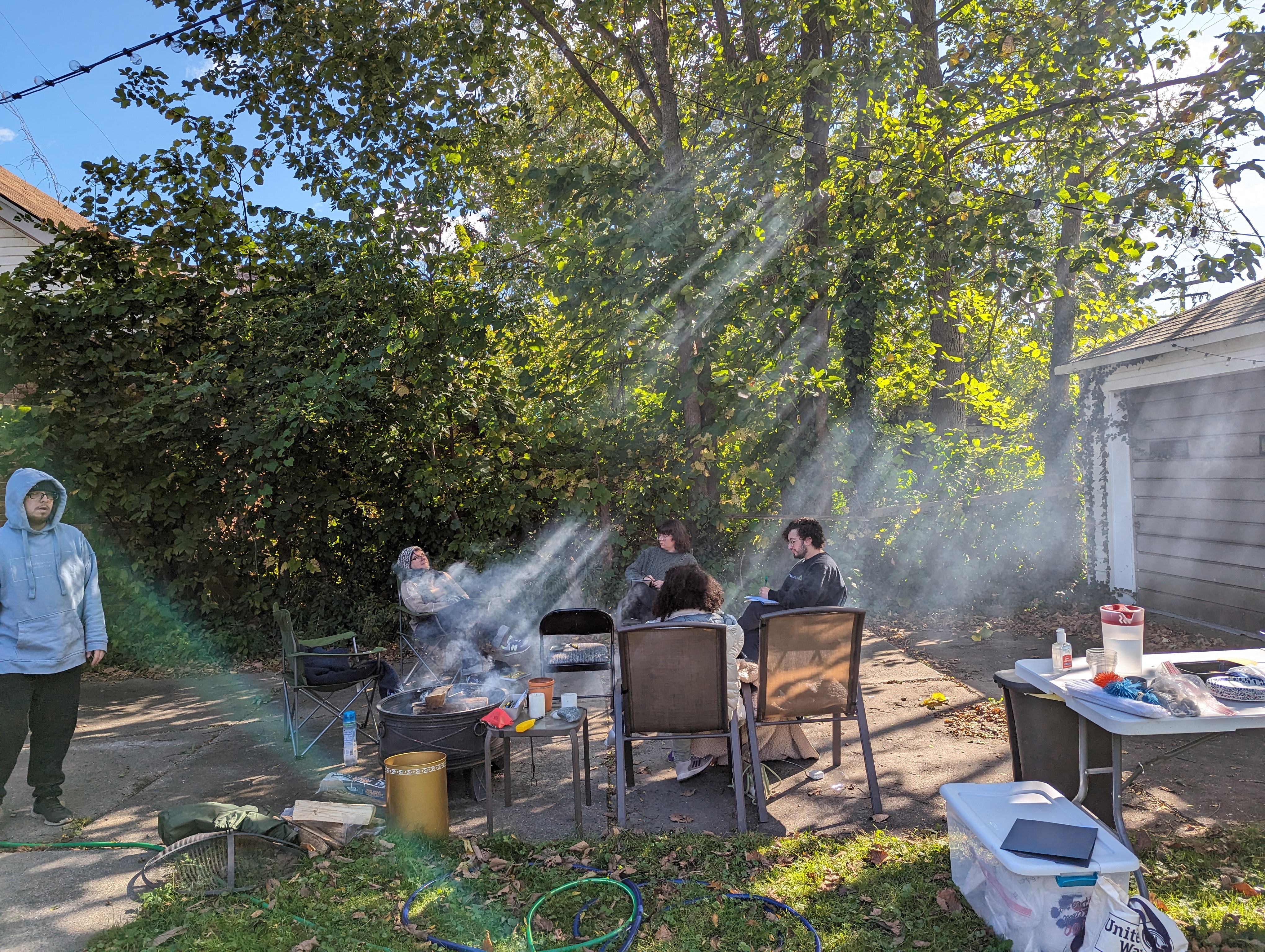 A photo of Detroit Peer Respite members sitting around a fire. The photo is taken from far away; the faces of individual members cannot be seen, but the image centers on the distinct rays of sun streaking dramatically through the trees and catching on the fire's smoke.