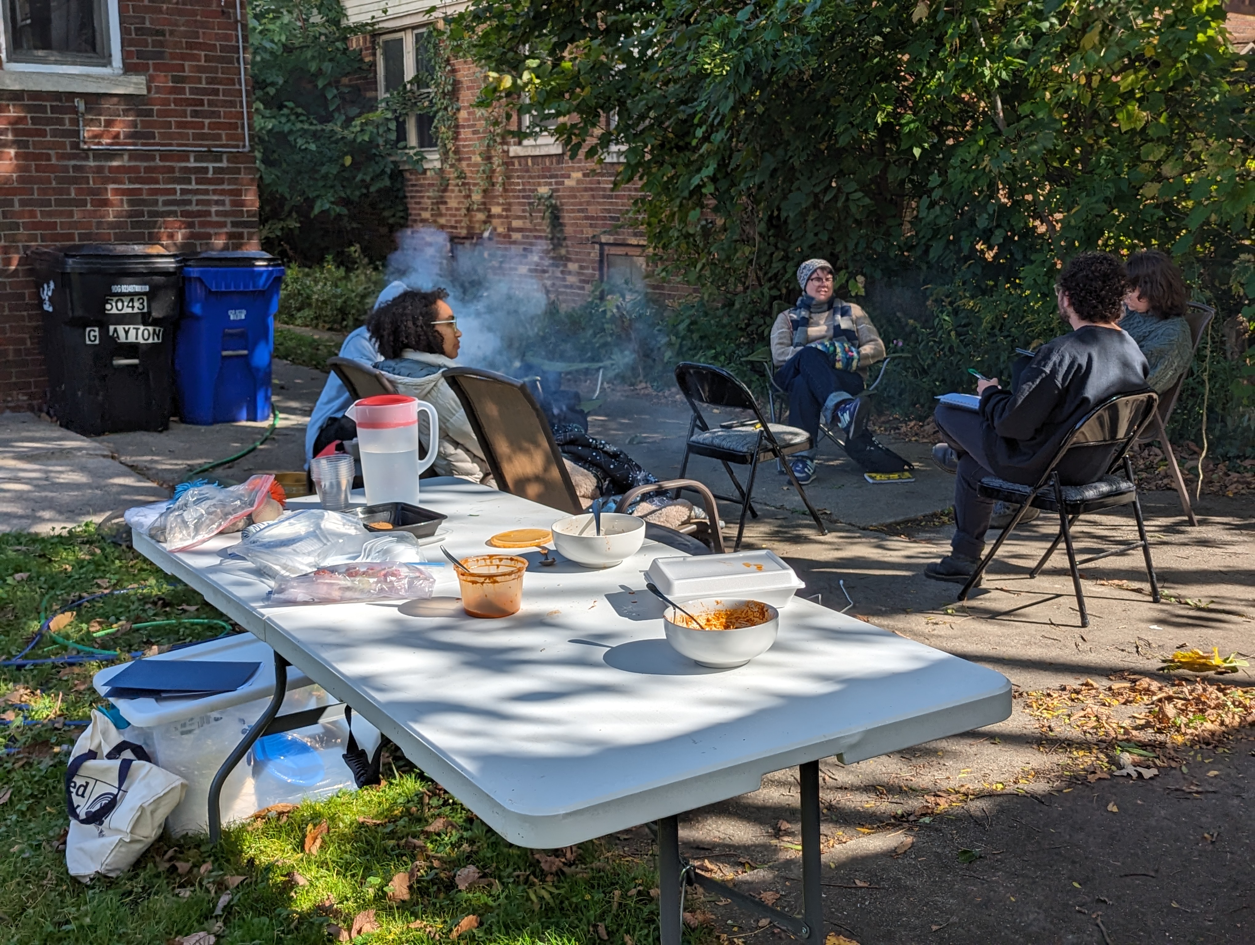 An image of Detroit Peer Respite members sitting in a circle around a fire. In the foreground is a large white folding table scattered with containers of food.